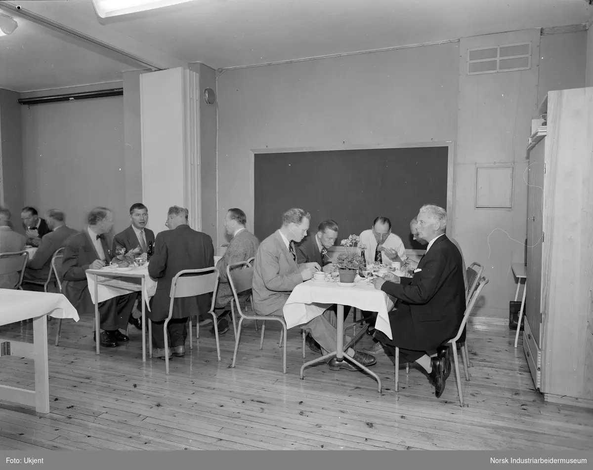 Interiør fra Velferden. Personer i pentøy samlet innendørs under måltid.