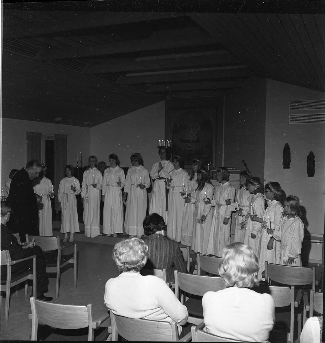 Ett luciatåg i Gränna står uppställda på rad framför en sittande publik. Lucian heter Anna-Beth Petri. Till vänster står kyrkoherde Wallis Wendel.