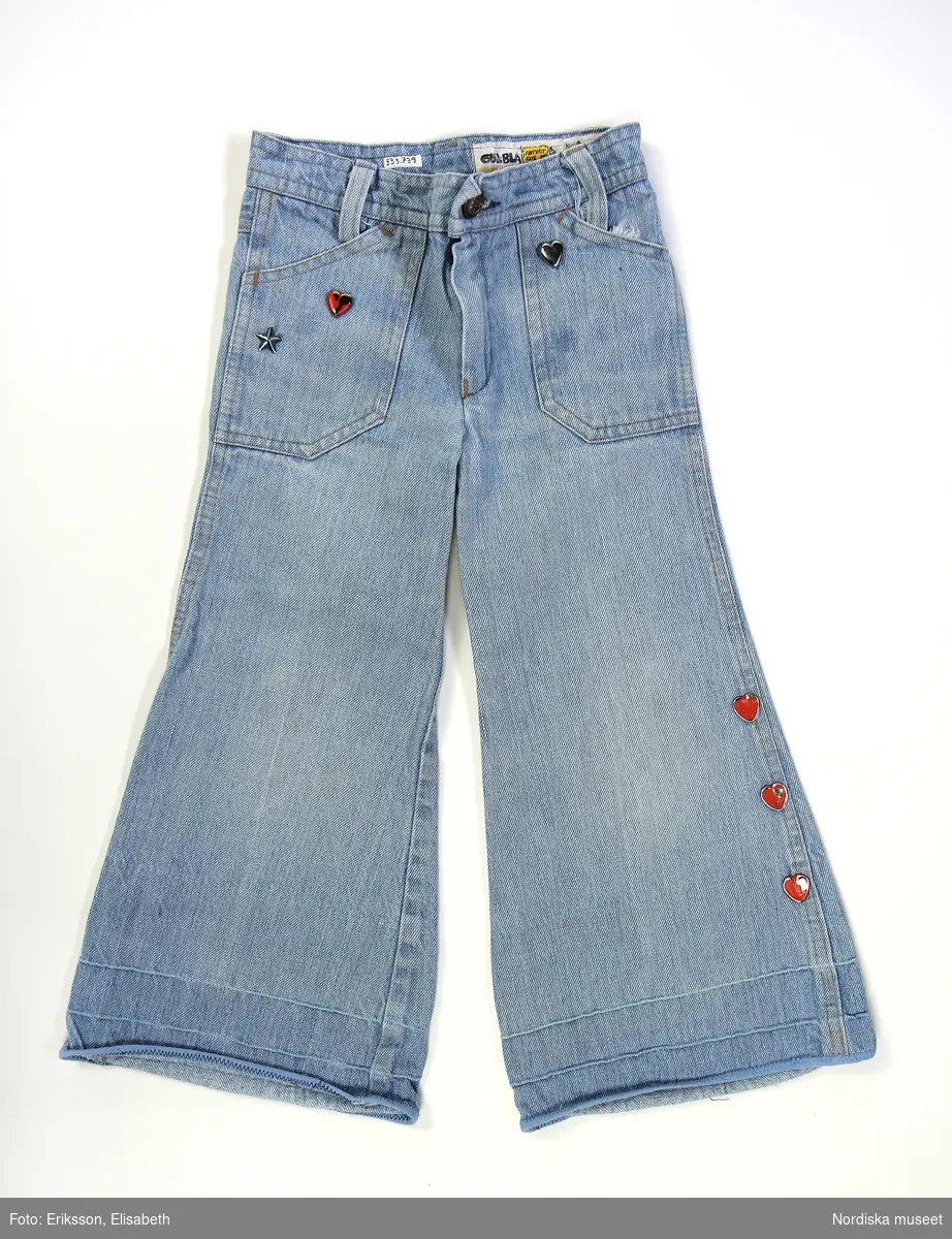 Utsvängda jeansbyxor för barn i 100 % bomull från Gul & Blå, barnstorlek 91. Metalldekorationer i form av stjärnor och hjärtan placerade vid fickor fram och bak samt vid nederkant längs med vänster sidsöm. 
2 framstycken och 2 bakstycken, 2 stora fickor framtill och 2 små fickor baktill. Besparing bak på jeansen. Alla sömmar är dubbelsöm, den klassiska så kallade jeanssömmen. Gylf av plast och metallknapp i midjan, av sorten patentknappar i mässing med stansat ”Gul & Blå”, samt en stjärna centrerat högst upp och i knappens nederkant. 3 hällor på bakstycke och 2 stycken fram. 

Etikett placerad på byxornas insida upptill med texten i pratbubblor från tecknade kaniner. Exempel på text: ” Gul & Blå får ej kopieras fintvätt 100 % bomull. Dra i byxan när den är fuktig.”  Nederkanten av byxbenen har två horisontala sömmar av ljusblått och mintgrönt, samt kanter med blått bomullsband. Byxan är nedlagd från originalsöm.
 
/Maria Vujovic 2018 & Hanna Leijon 2019-03-08