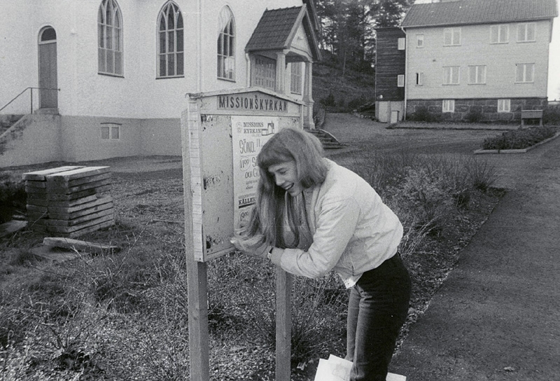 En okänd ung kvinna lutar sig framåt och skrattar, okänt årtal. Hon står framför en skylt tillhörande Kållereds missionsförsamling. Till vänster ses Kållereds Missionskyrkan (senare Ekenkyrkan) och i bakgrunden till höger ses före detta Villa Fridhem, som nu är lägenheter.