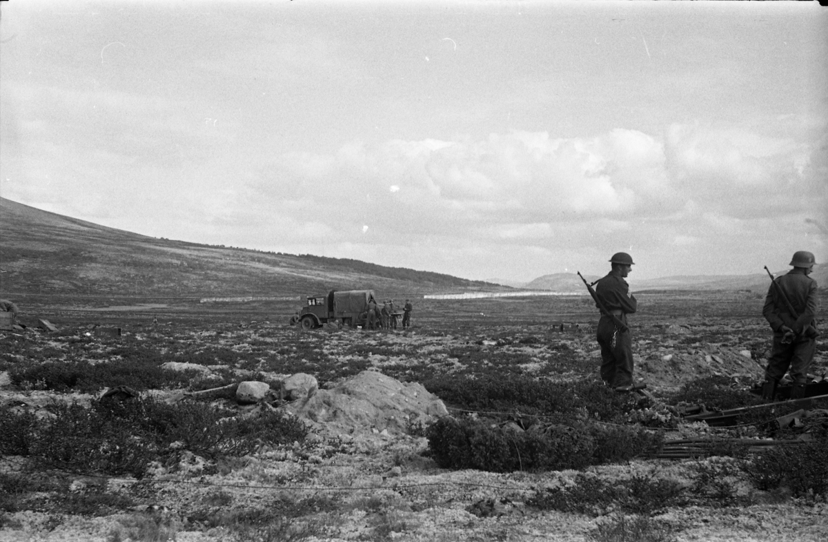 Avfotografert landskapsbilde med to soldater i forgrunnen til høyre i bildet. Midt i bildet et militært kjøretøy og flere seoldater. Hverken personer eller sted er identifisert.