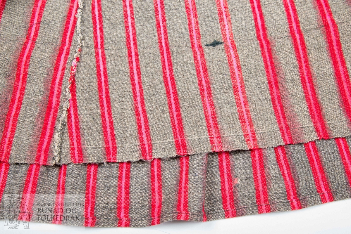 Stripet halvullstakk med grå botn og striper i rosa og raudt.  Kappe nede og skrådde breidder med to folder ved splitt midt bak. Knyteband ved splitten. H.: 78 cm.