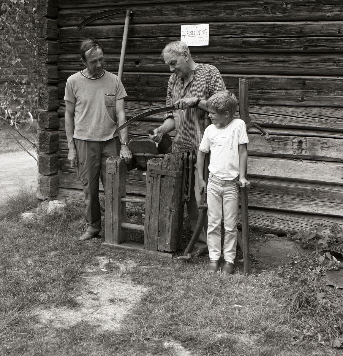 Två män och ett barn står intill en husvägg. Den ena mannen håller i ett blad till en lie och slipar detta under Rengsjöfesten den 14 juli 1985.