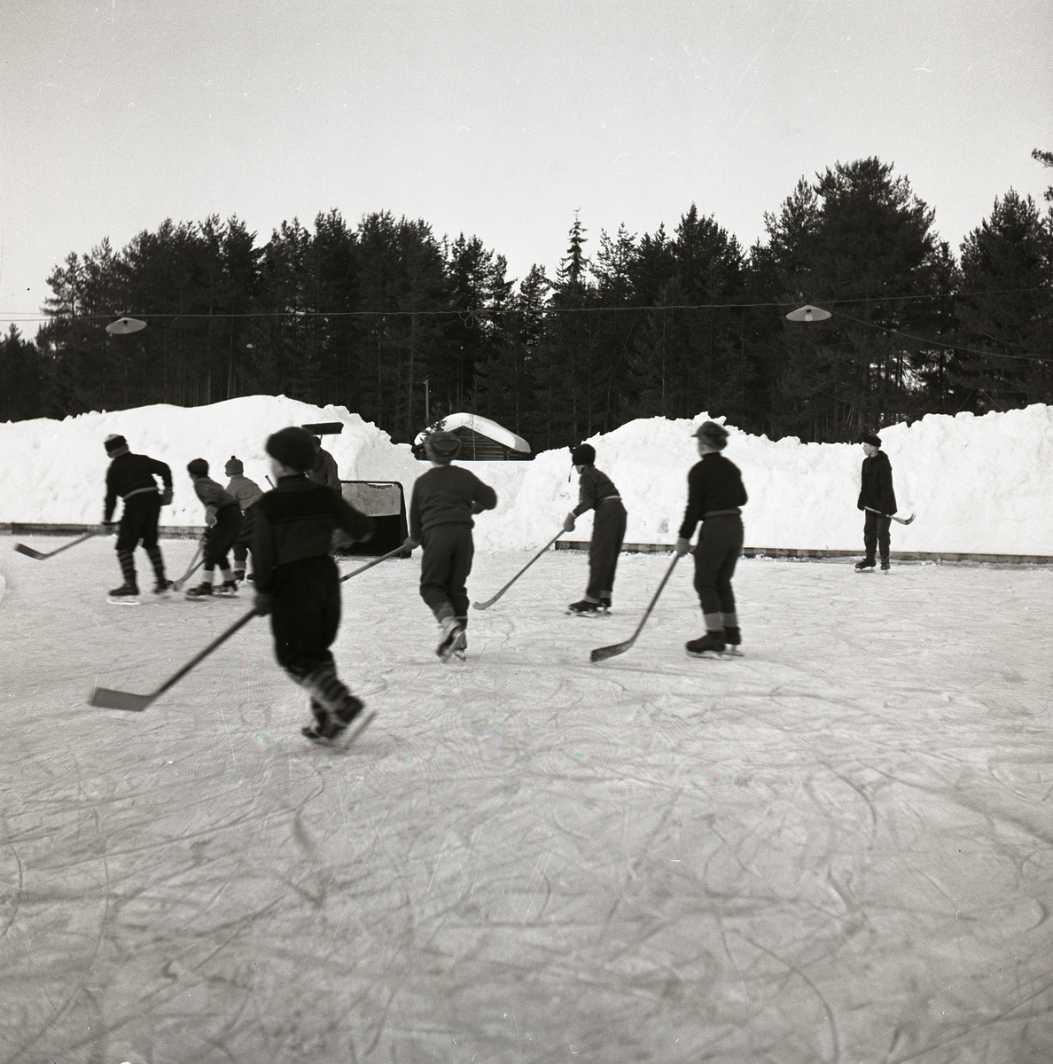 En grupp med pojkar spelar ishockey på isplanen dom har skridskor och ishockeyklubbor. Det finns målburar, snöhögar, byggnad, skog och himmel. Vid isplanen finns det en sarg och belysning ovanför. Ishockey Glösboplanen 27 januari 1959.