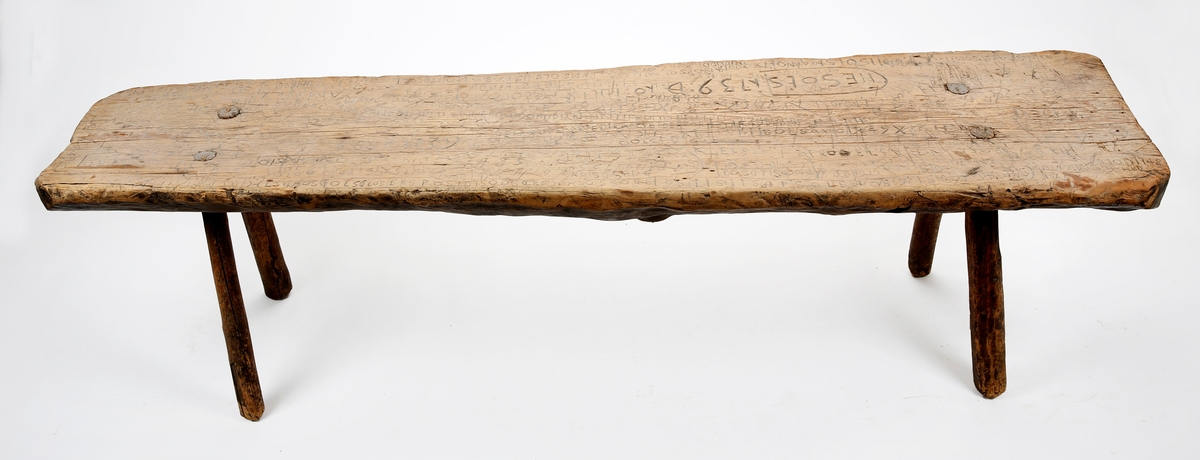 Enkel, grovt utformet benk med fire naturvokste ben. Sitteflaten er dekket av utskårne bokstaver og tall.