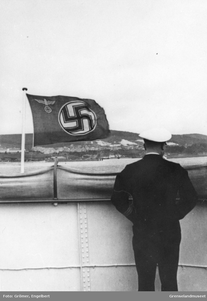 Skipsoffiser står akterut på et skip som går fra land. Flagg med hakekors som motiv med tysk ørn stående på hakekors i øvre hjørne ved festeanordning. 