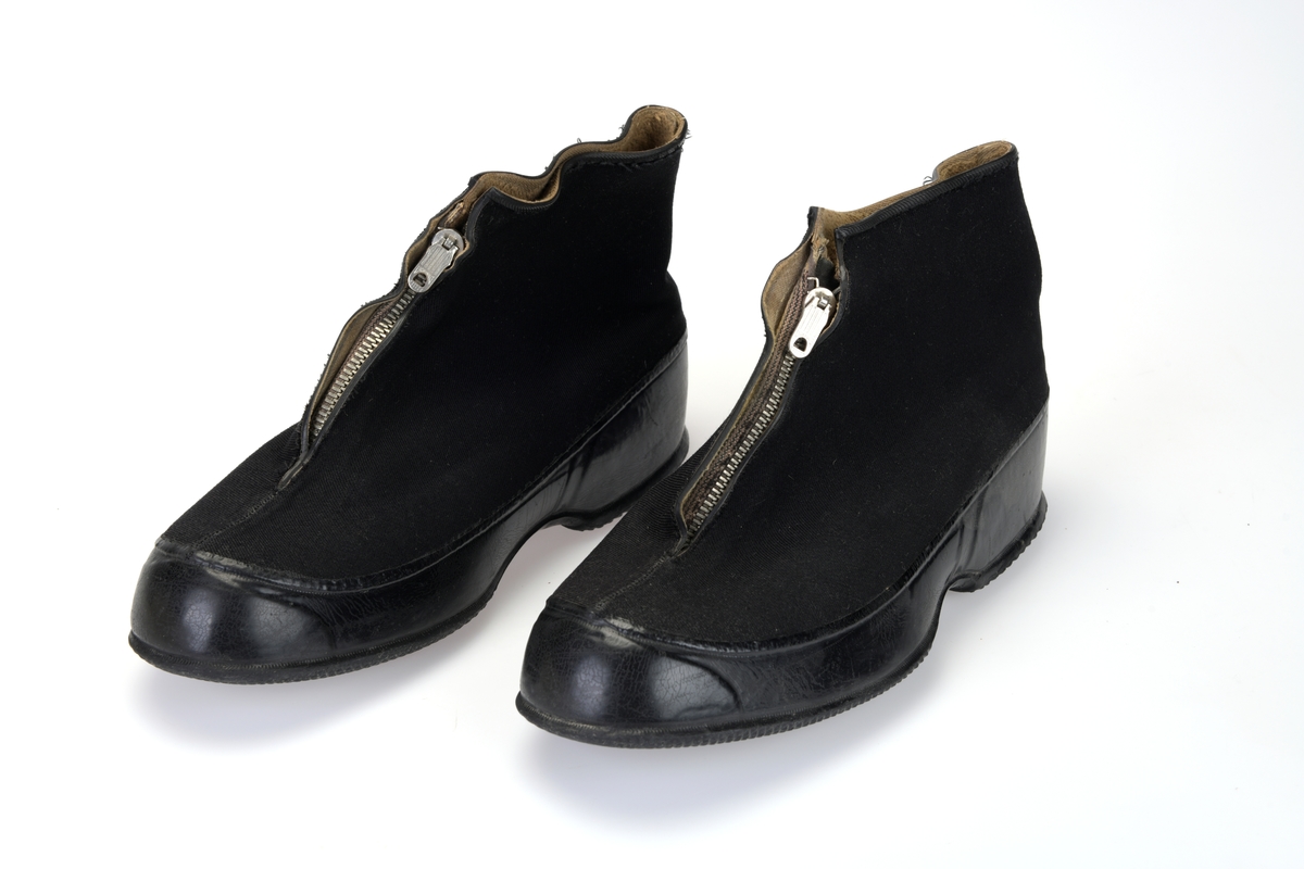 Et par svarte herresko i skoeske. Det er overtrekkssko/kalosjer. Overdelen er laget av svart bomullsstoff. Innvendig er det lysebrunt bomullsstoff. I front er det en 13cm lang glidelås. Bak glidelåsen er det svart bomullsstoff som forsterkning mot fukt. Langs kanten oppe og langs glidelåsen er det en smal gummikant. Nede på skoen er det en gummikant som er 5,5cm på det bredeste. Sålen er laget av gummi og den er smårutet. På undersiden av sålen er det påført tekst, se "Påført tekst/merker". Skoene ligger i en skoeske av hvit papp. Esken er stfitet sammen. På den ene kortsiden er det informasjon om produsenten, se "Påført tekst/merker".
