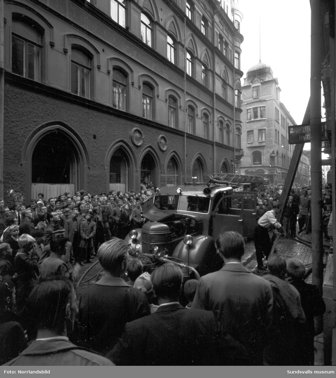 En stor folksamling ser på då brandkåren bekämpar en brand på Nybrogatan 8, En stege är rest invid kaffeaffären Ido.