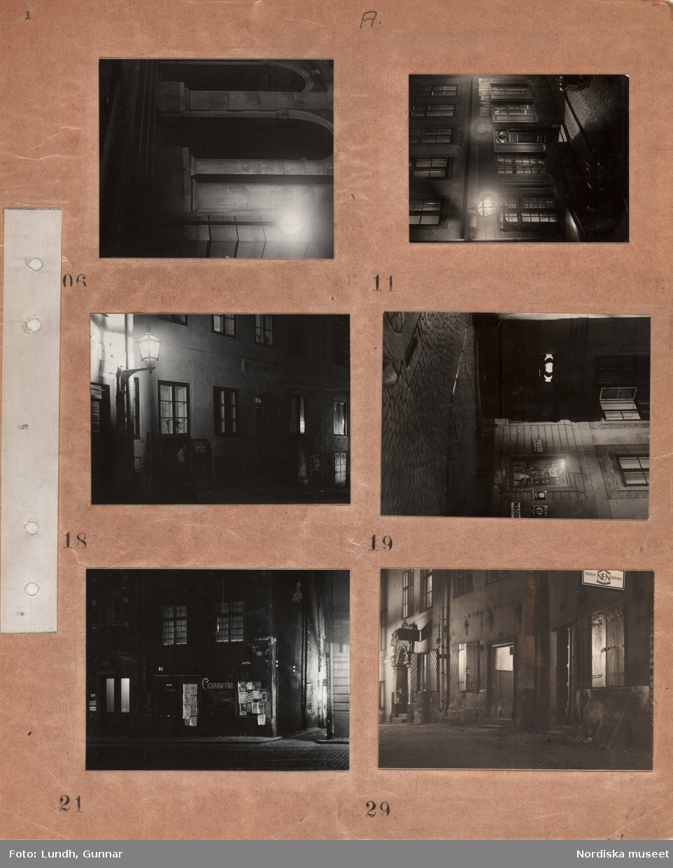 Motiv: (ingen anteckning) ;
Nattbild av stad med gator och byggnader möjligen Gamla Stan, husfasader med upplysta fönster, skyltfönster med skylt "Frisör" "Tvättbjörn", hus med skylt "Cigaraffär" och löpsedlar och affischer.