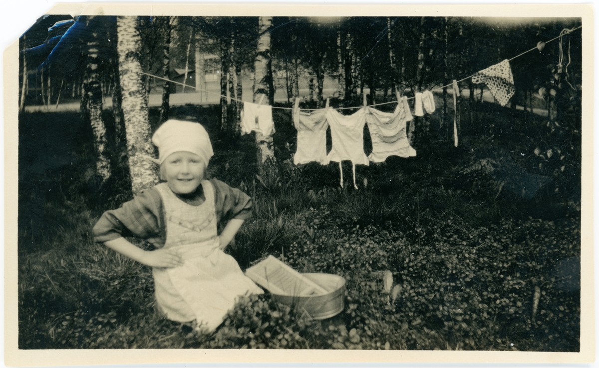 Barn som sitter bredvid tvättbräda och balja. I bakgrunden hänger dockkläder på en tvättlina
