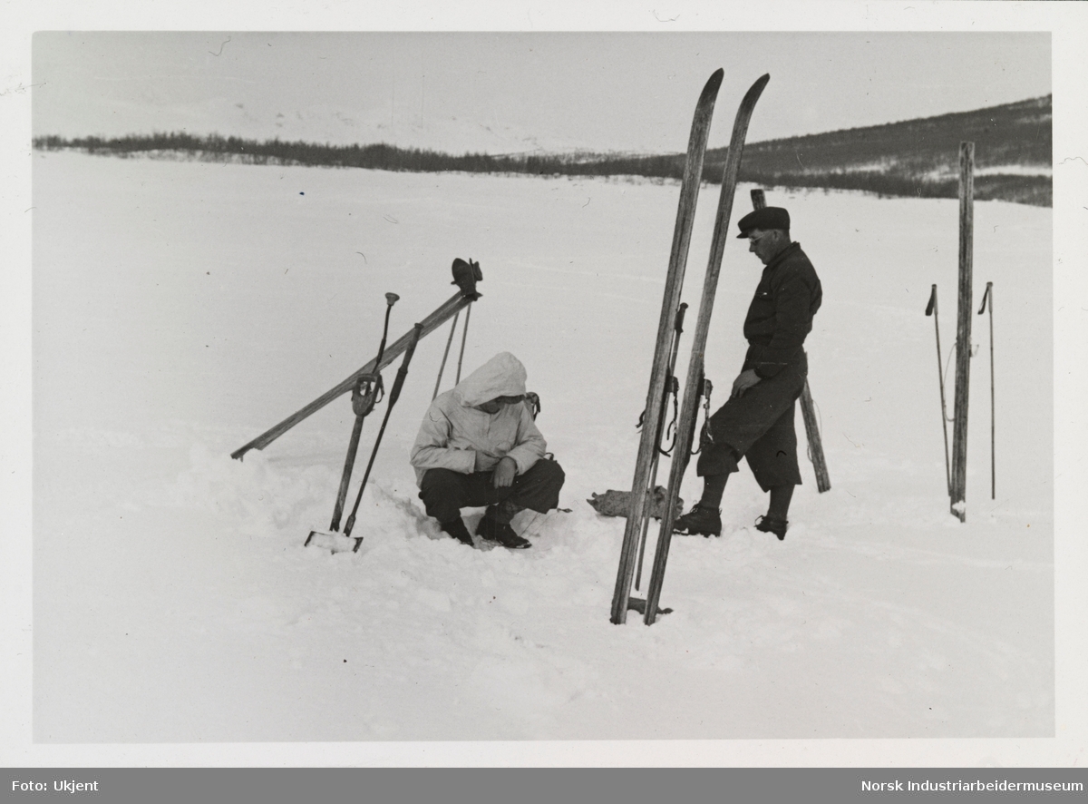 Mann sitter og isfisker, ved siden står en mann og rundt er ski og skistaver plassert i snøen