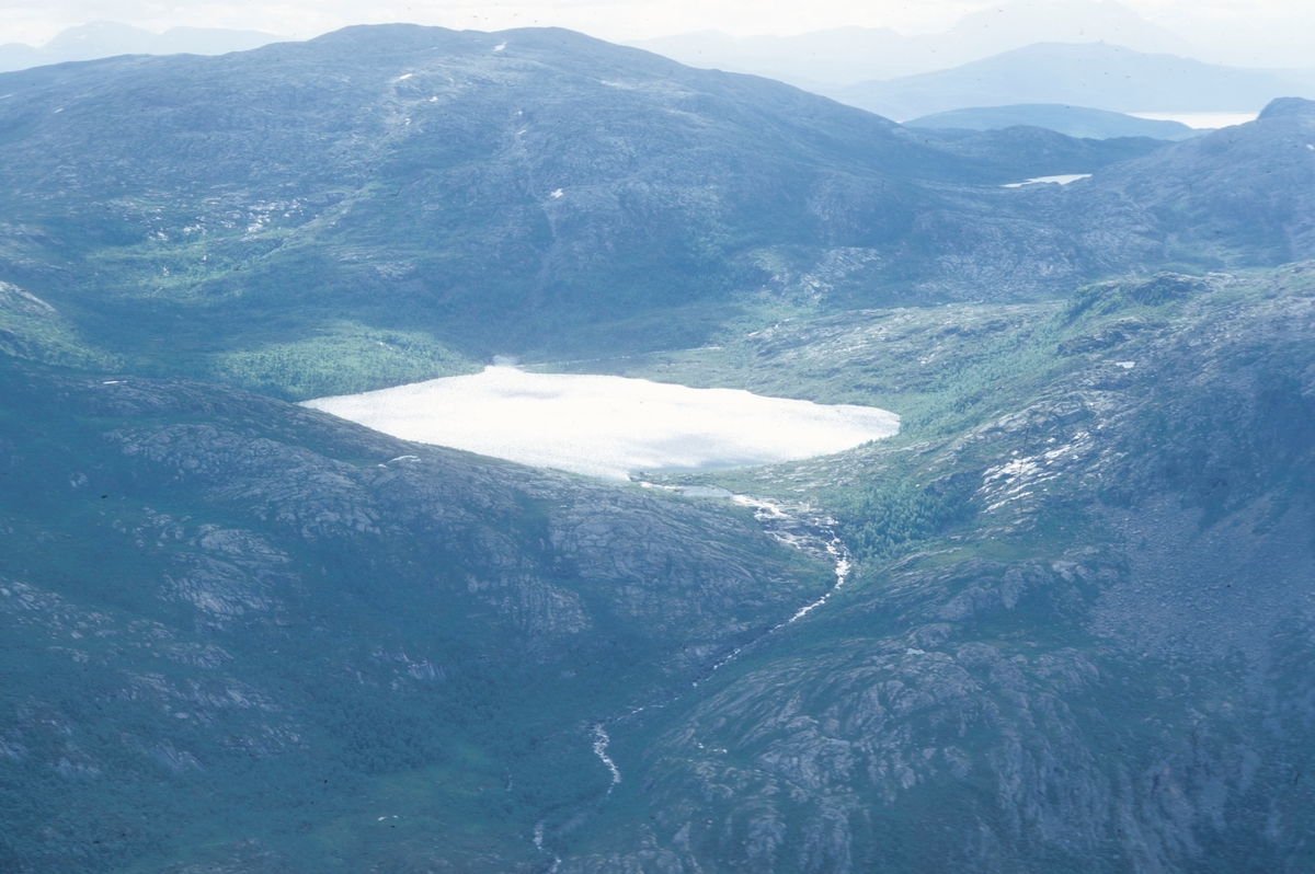 Flyfoto av et vann, omsluttet av fjell. I forgrunnen renner ei lita elv fra vannet.