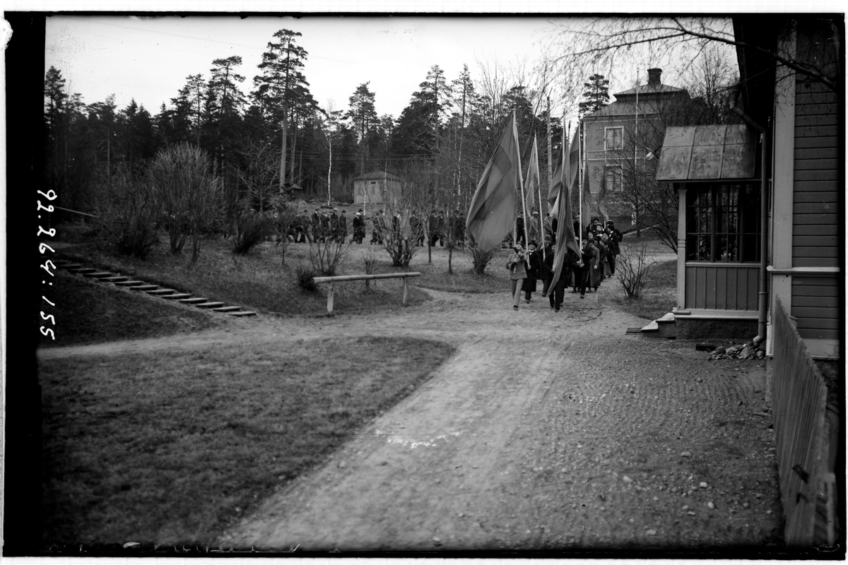 Hålahult sanatorium, exteriör, många människor marscherar i samlad trupp, fyra fanbärare först, på sjukhus-området.