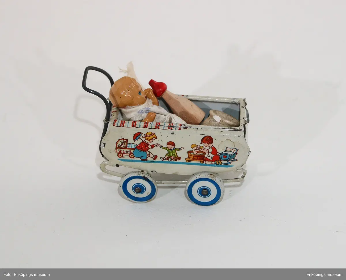 Dockvagn av plåt med 4 hjul. På sidorna har den motiv av lekande barn. I vagnen finns en docka nerbäddad. Finns en keramikpotta.Vet inte om de tillhör.