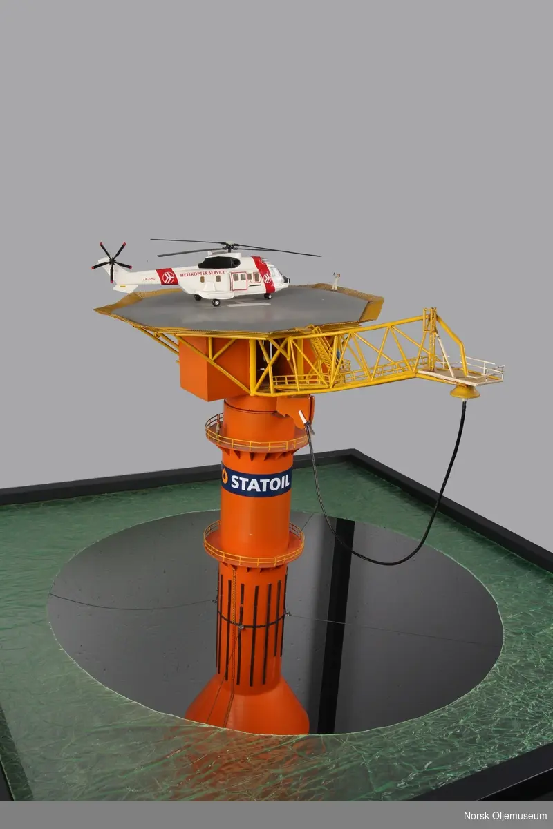 Modell av lastebøyen knyttet til Statfjord A. Malt orange og gul med helikopter på helidekket. Modellen holdes på plass av en aluminiumsramme der bunn og topp forestiller hhv. havbunn og hav.