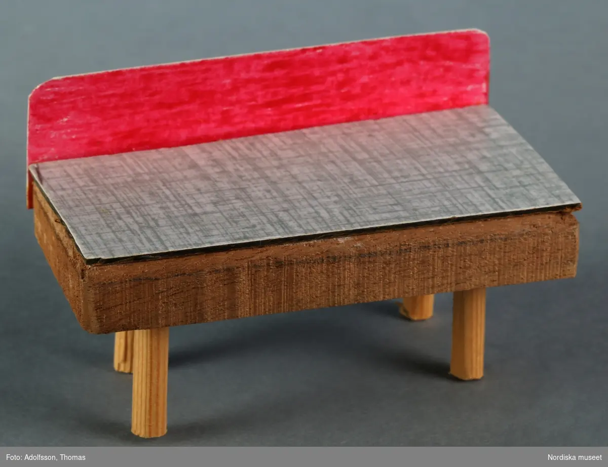 Ett hemtillverkat bord av trä och rödmålat bakstycke av kartong, med skiva av perstorpsplatta. Hör till dockskåpsinredningen i källarens hobbyrum i dockskåp NM.0331721+.