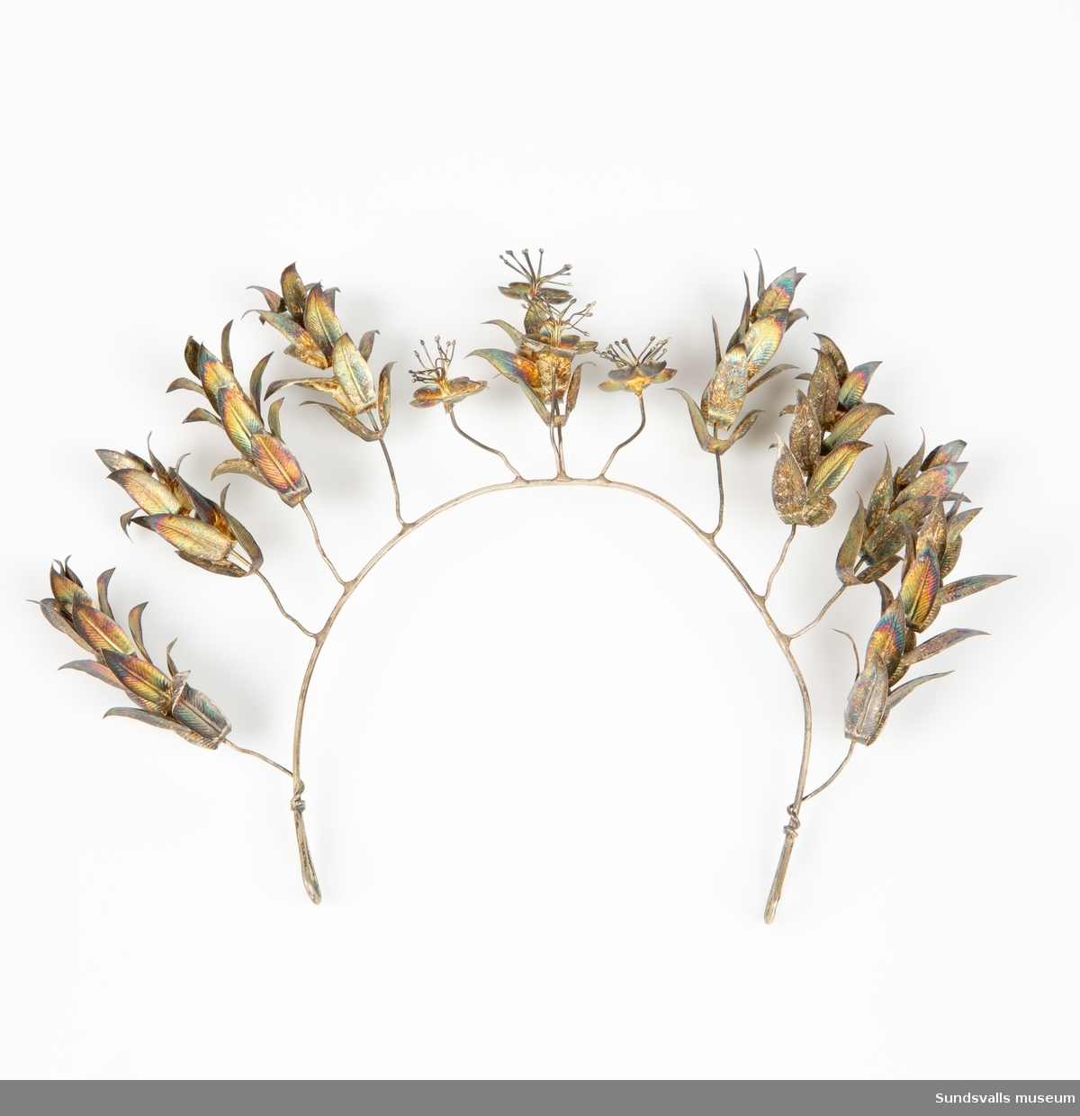 Diadem i böjd metalltråd med en ögla i var ände. Dekoration i form av metalltrådar med blommor och blad är fästade runt den böjda tråden.