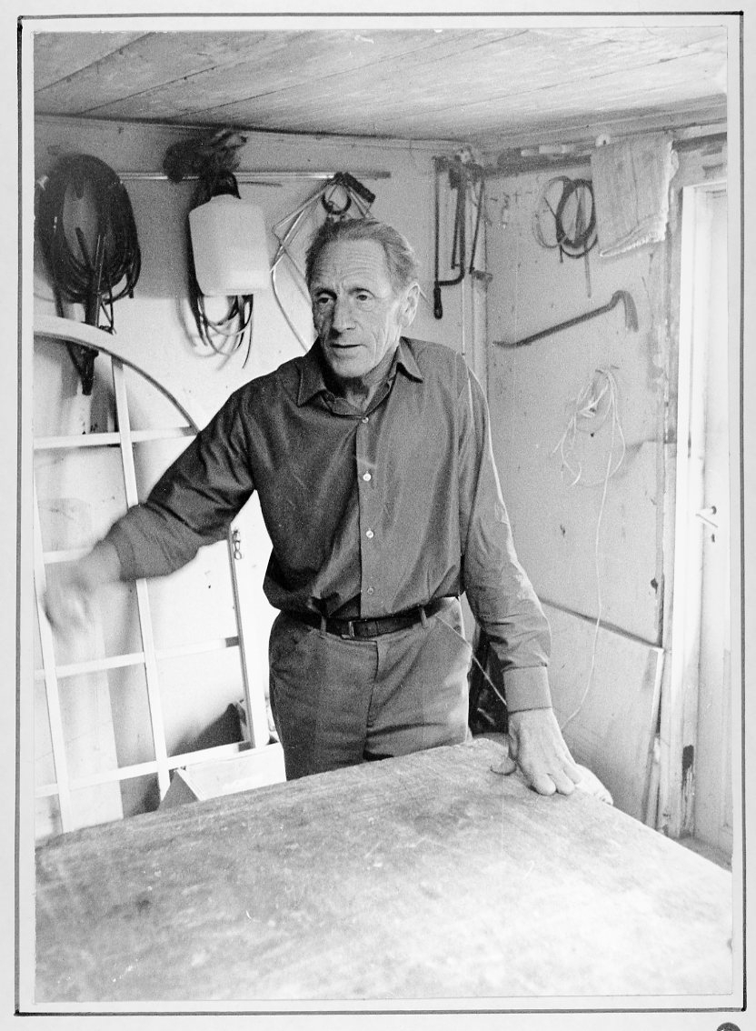 Porträtt av glasmästare Bernt Rendahl i glasmästeriverkstaden. Han står lutad mot ett bord, gestikulerar med höger hand.
