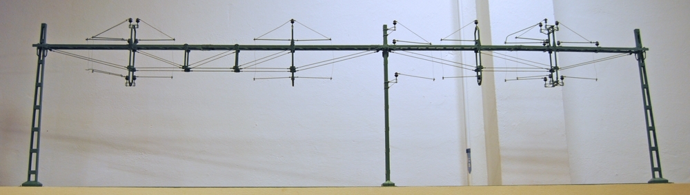 Modell i skala 1:10 av kontaktledningsbrygga, fackverksbrygga med tre bryggstolpar för åtta spår. Grönmålad med bruna isolatorer.