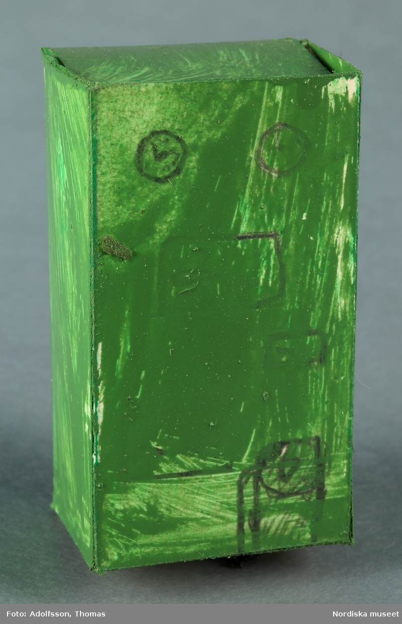En oljepanna av ihopvikt pappkartong, bemålad med grön färg. Mätare och andra detaljer är påritade med blyerts eller bläck. Oljepannan hör till inredningen i källarens pannrum i dockskåpet NM.0331721+.