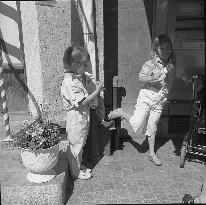 En pojke och en flicka äter polkagris. De står delvis i skugga intill en barnvagn och en fastighet med en bankomat.