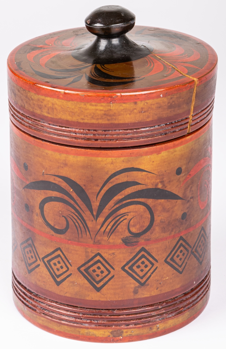Kat.kort:
Tobaksburk av trä, svarvad, med knoppförsett lock, cylindrisk. Gulbrun bottenfärg med dekor i svart och rött.