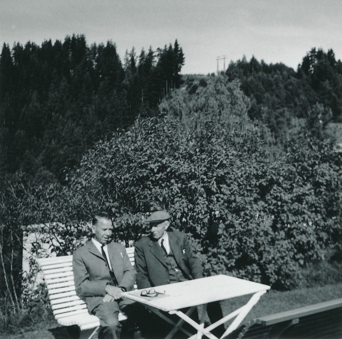 2 menn i en hage sitter på benk med bord foran seg.