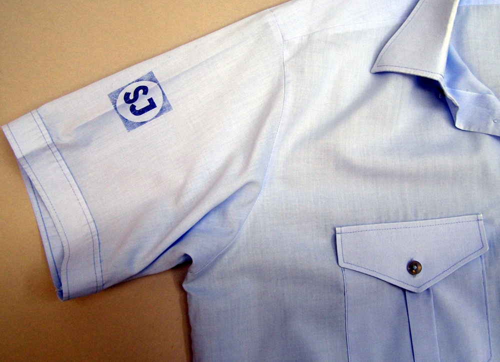 Kortärmad skjorta av tunnt ljusblått bomullstyg med ok, krage och två bröstfickor med lock och knäppning framtill. Skjortan knäpps fram med små knappar av grå-vit plast. Märkning/tryck i mörkt blått på höger ärm ovantill: SJ:s logga i fyrkant inom en cirkel, det såkallade "frimärket".
Skjortan är ej märkt med storlek.