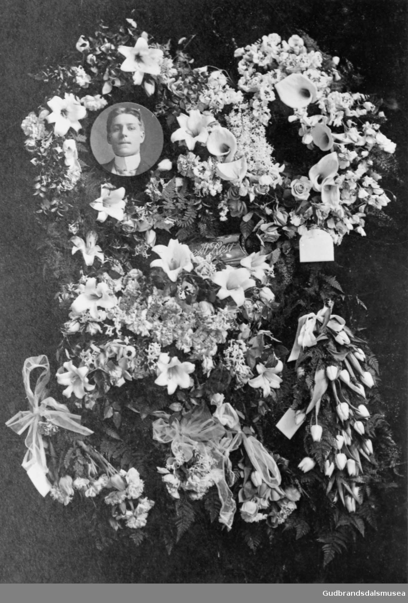 Lite portrettbilde av Syver Enebo er plassert i en blomsteroppsats, mange blomsterkranser rundt. Til minne om?