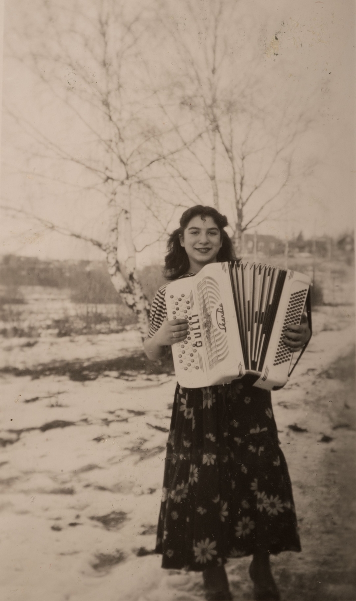 En ung romsk kvinna spelar dragspel. Runt om henne syns ett vinterlandskap med snö och kala björkar.