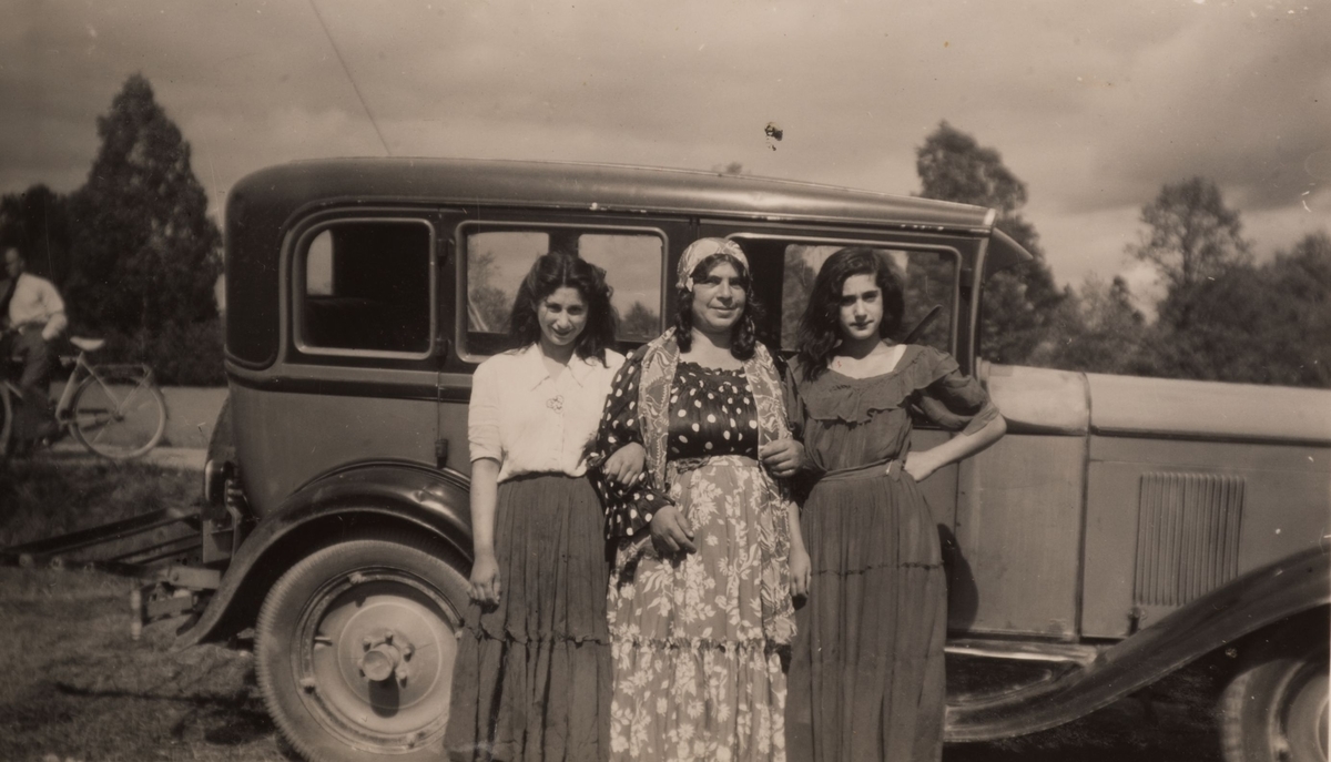 Tre romska kvinnor framför en bil i Sandviken under sommaren 1947. I bakgrunden syns en man lutad mot sin cykel.