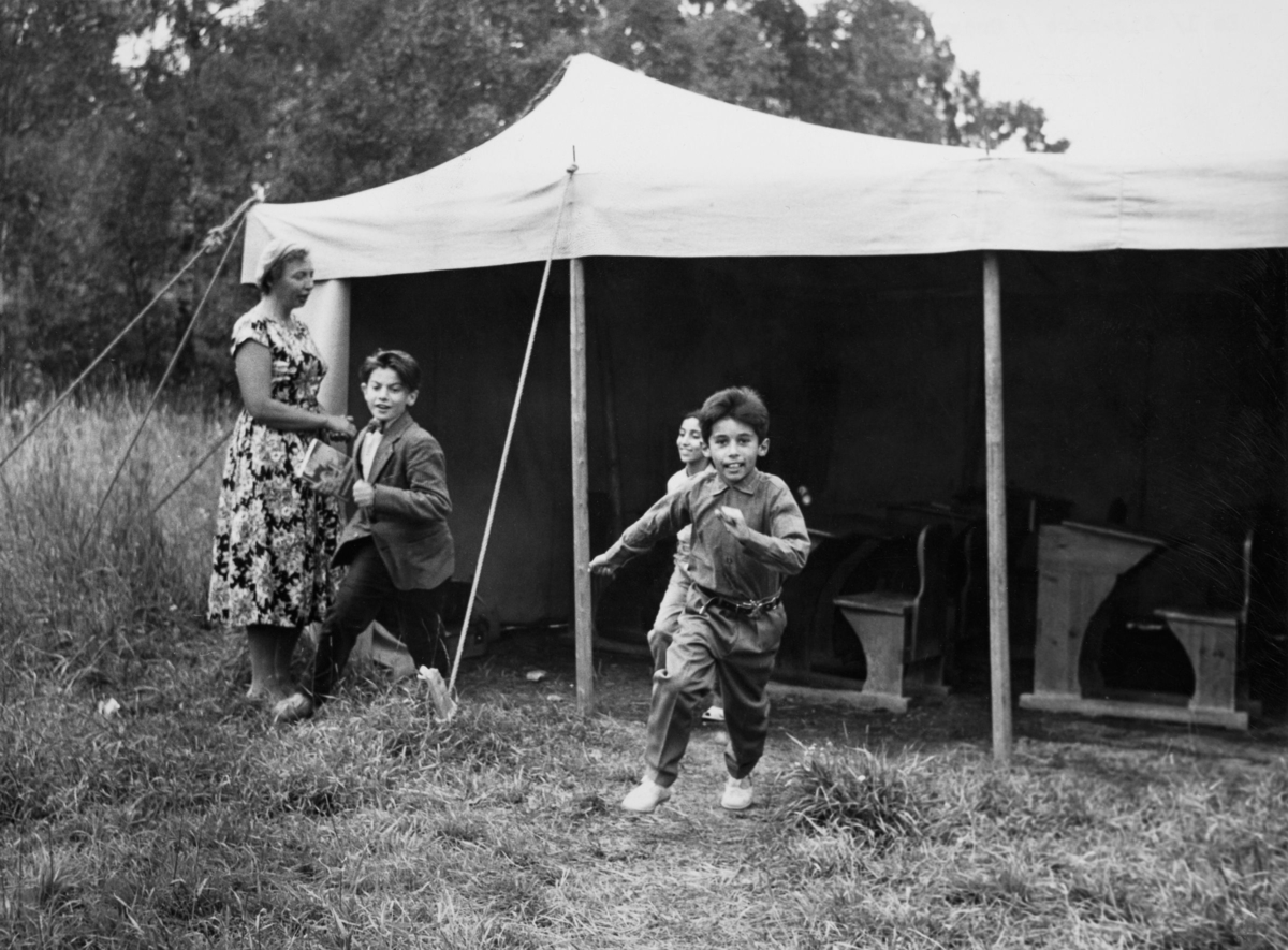 Tre romska barn springer ur från skoltältet där fröken står kvar. I tältet står skolbänkar.