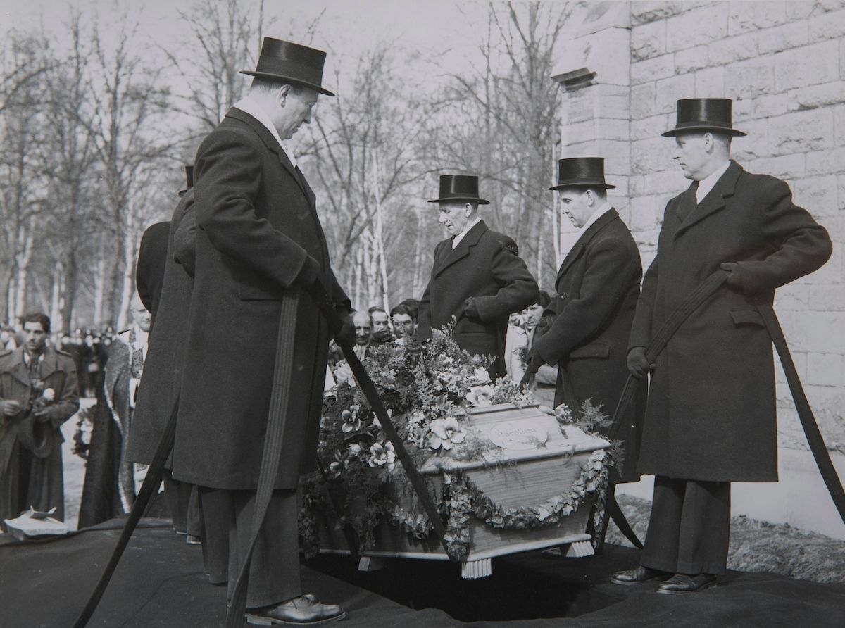 En kista sänks med i en grav av herrar iförda rockar och svarta hattar. I bakgrunden syns en kyrka.