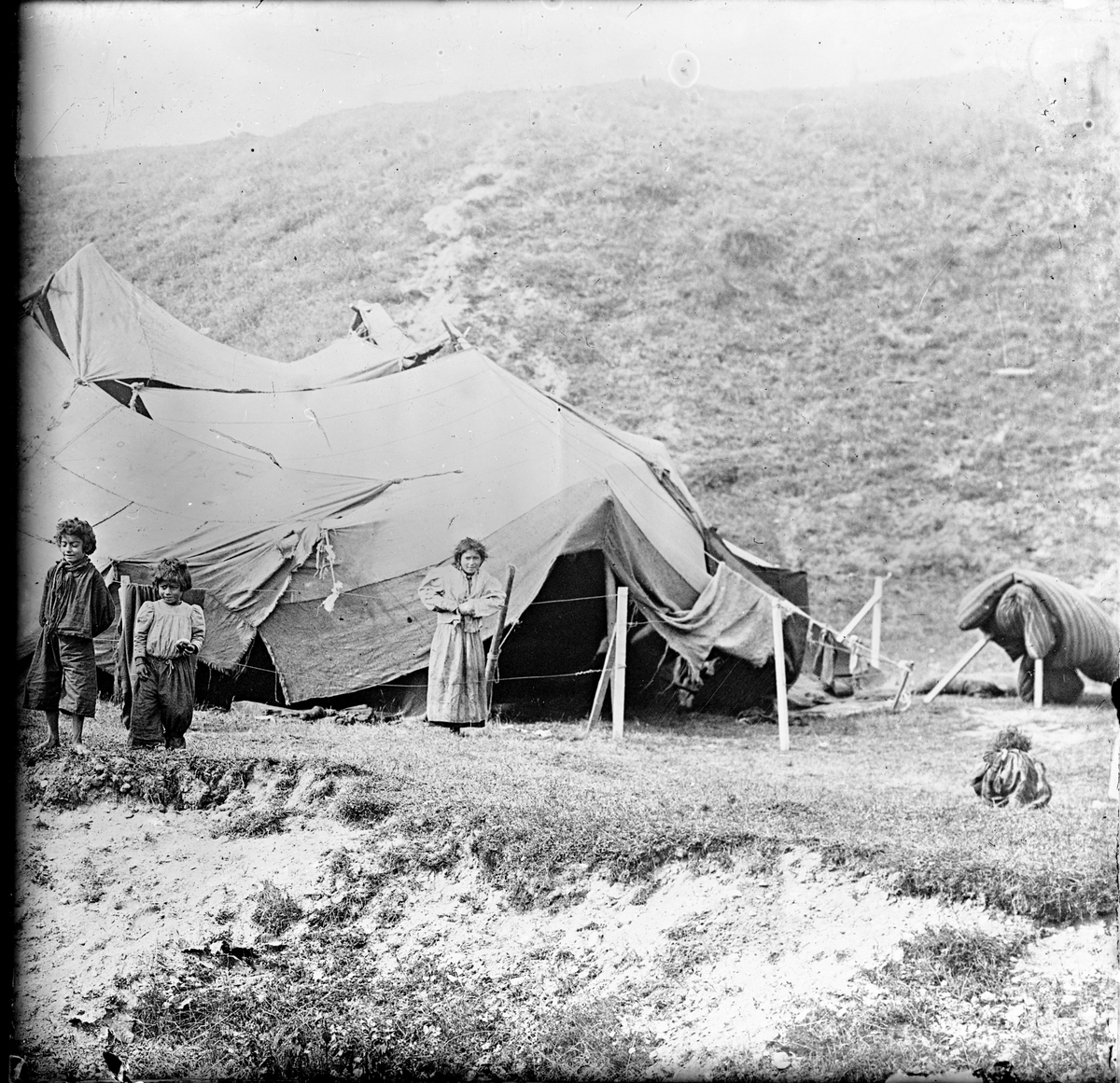 I en backe har ett romskt sällskap slagit läger. Ett tält syns i bakgrunden och framför det tre barn i olika åldrar.