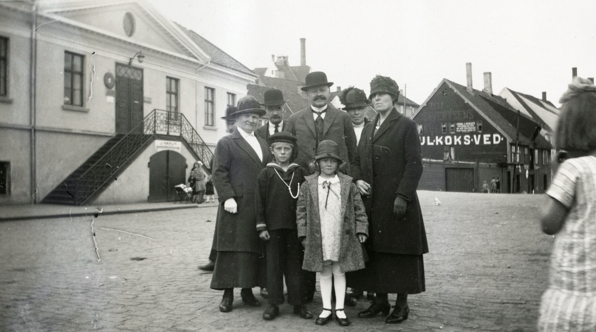Gruppeportrett av en familie på gata i en by. I bakgrunnen er en bygning med skriften kul, koks, ved. På den andre står det J. Nærum (dampskipekspedisjon?)
