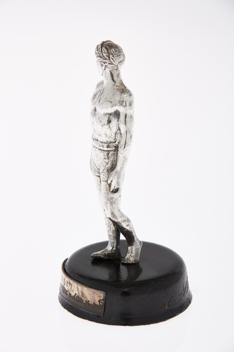 Statuett i sølvfarget metall, en atlet, med støtte i tre. Plakett på trestøtten med inskripsjon.