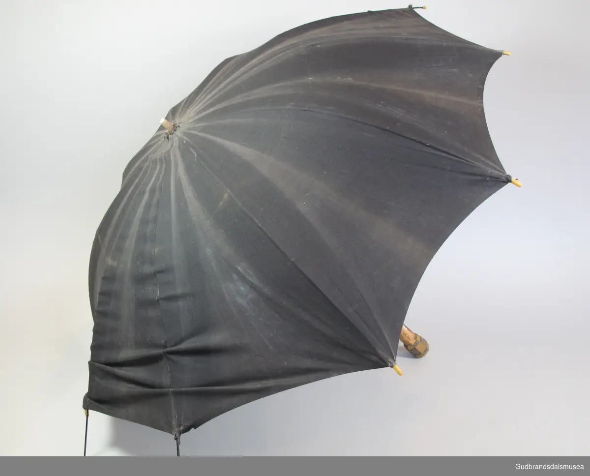 Liten dameparaply fra 1930-50-tallet. Skjerm i svart stoff. Ottekantet håndtak i tre belagt med plastlameller. Spilere i metall. Stang i tre. Liten lærstropp for omvikling av paraply i sammenslått tilstand.