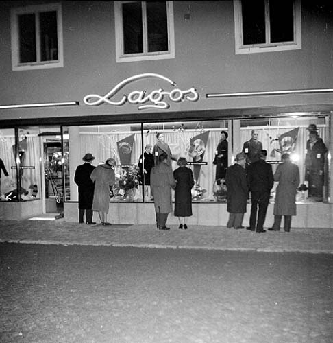 Kvinnor och män, i armkrok, i rock och hatt står och tittar i upplysta skyltfönster till butiken Lagos (Valte Lago Dam och Herrekipering) på Brahegatan i Gränna.