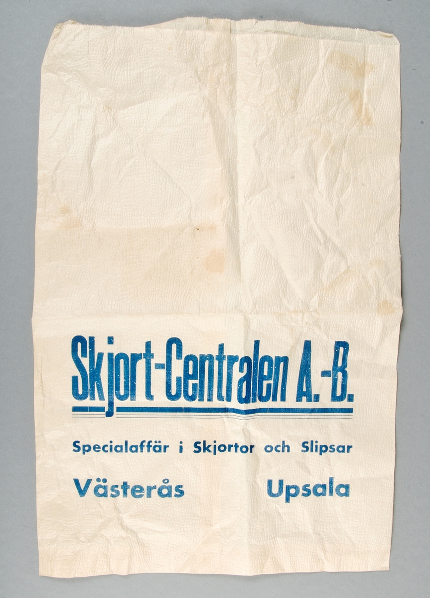 Gulbrun papperspåse. Lite räfflad struktur. Text i blått: Skjort-Centralen A.-B. Specialaffär i Skjortor och Slipsar. Västerås Upsala.