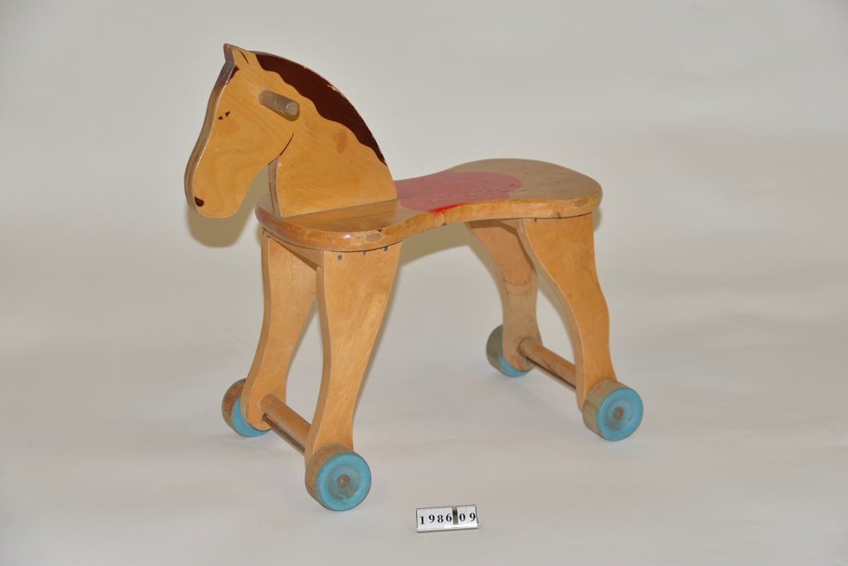 Pallhäst på hjul, tillverkad av massivt trä. Röd sadel, brun man och blå hjul, i övrigt naturfärgad.