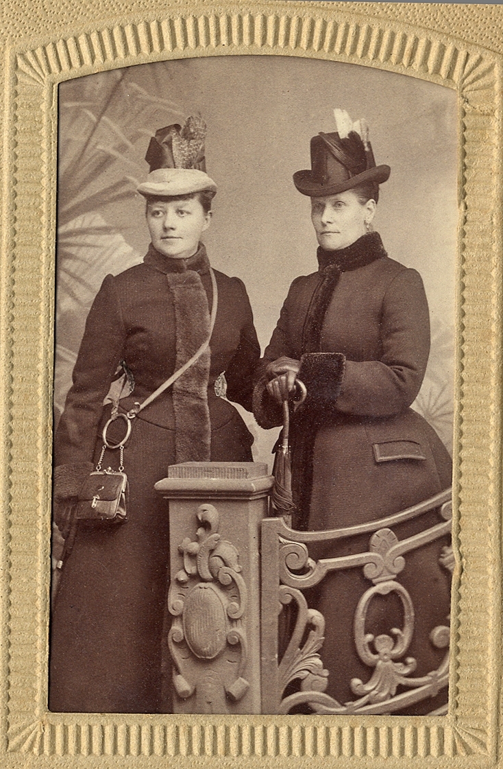 Två kvinnor i vinterkläder (kappa, hatt och handskar). 
Ateljéfoto. Knäbild, halvprofil.