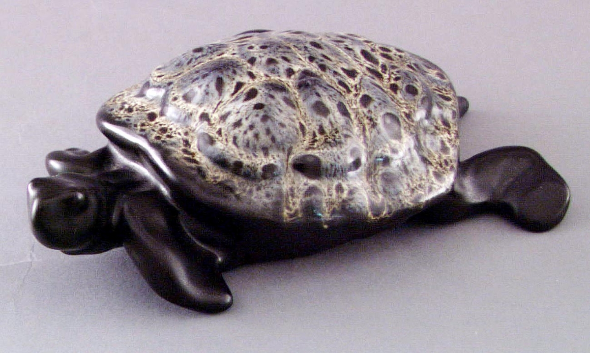 Figurinen Sköldpadda av formpressad flintgods med svart glasyr, ryggen gul- och gråmelerad. Formgiven av konstnär Lillemor Mannerheim 1955.
