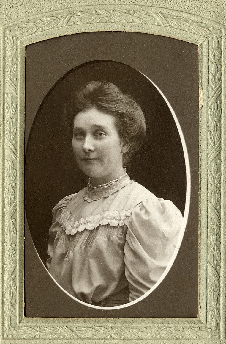 En okänd ung kvinna i ljus höghalsad klänning med stråveck och tättsittande pärlhalsband. 
Bröstbild, halvprofil. Ateljéfoto.