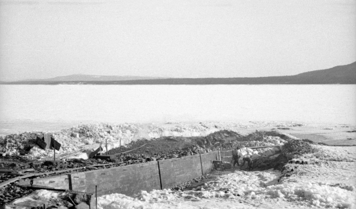 I 1939 kjøpte Glomma fellesfløtingsforeningen eiendommen Sørlistøa på vestsida av Osensjøen i Åmot.  Her ble det bygd et senter for fløtinga på sjøen med hytte som inneholdt kontor for fløtersjefen og mannskapsrom, båthus, vedskur og smia.  For at D/S Trysil-Knut skulle kunne trekkes på land hver vinter bygde en vinteren og våren 1941 - da vannstanden i sjøen var lav - en bedding.  Dette fotografiet er tatt i forbindelse med dette arbeidet.  Bildet viser en betongmur som går nedover mot sjøen.  Aller nederst er noe av forskalinga fortsatt intakt.  Sjøen er fortsatt islagt og snødekt, men på baksida av betongmuren er det opplagt en del masse som ikke er snødekt. 