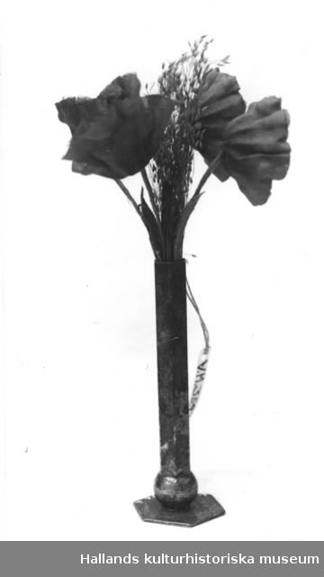 Dockskåpsvas med blommor. Nysilvervas, sex-kantig, längst ned en kulformad del och en sex-kantig fot. Tygblommor i grönt och rött. Blommornas längd: 10 cm.
