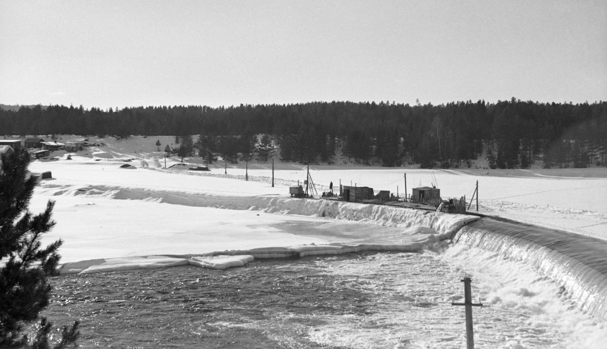 Anleggsarbeid på den midtre delen av den drøyt 300 meter lange terskeldammen ved Skjefstadfossen i Heradsbygda i Elverum, Hedmark. Fotografiet ble tatt fra østre elvebredd i slutten av februar måned i 1942. Da var elva, med unntak av det østre løpet nedenfor dammen (i forgrunnen, der vannføringa var størst) is- og snødekt. Anleggsarbeiderne hadde rigget seg til med brakker ved den midtre delen av dammen, og de hadde fått strømforsyning og oppkjørt veg ut til arbeidsstedet. Sannsynligvis skulle de forsterke dammen, som på dette tidspunktet var drøyt 30 år gammel. Slikt arbeid ble gjerne gjort i vintesesongen, for da var vannføringa i elva liten. Til venstre i bildet, bak og under noen bladløse kvister, skimter vi sjølve kraftverksdammen og tilhørende bygninger ved vestre elvebredd.