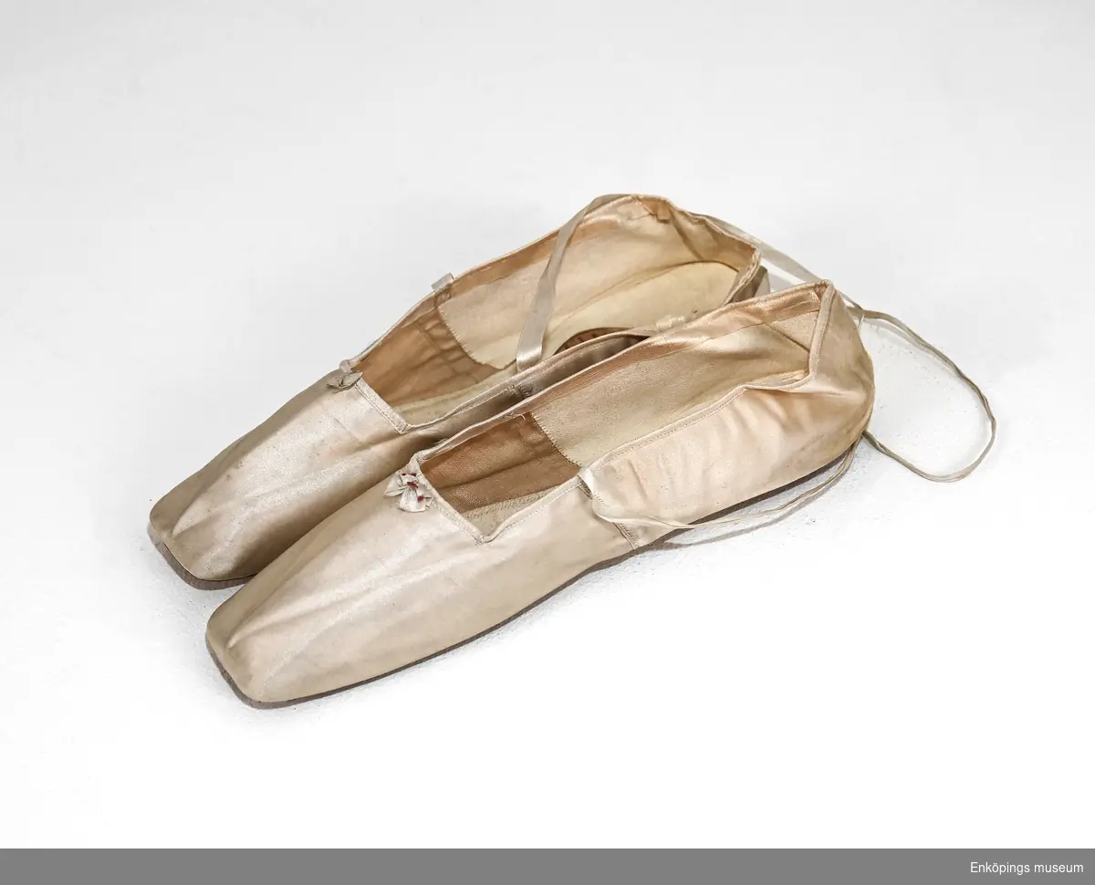 Cremefärgade balettskor med skinnfoder. Försedd med sidenband för att kunna knyta skorna runt vristen och en liten sidenblomma som dekoration. 
Tillverkare: Dufossée Müller et Melnotte, Paris, Frankrike.
Troligtvis från mitten av 1800-talet.