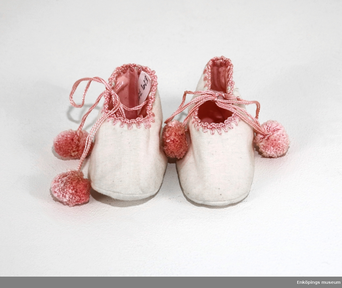 Vita flanellskor med rosa foder, toffelmodell. Skorna har virkad kant i rosa och knytband med bolltofsar. Tofflorna är tillverkade 1922.