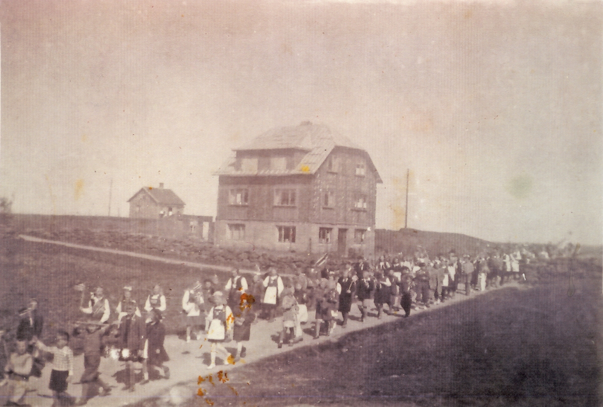 Barn med bunader og ellers finklær går langs en vei i 17.mai tog. Større hus midt i bildet. Hus i bakgrunnen.