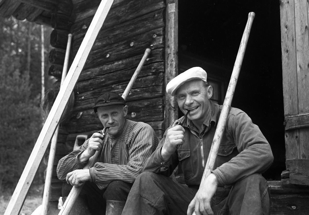 Ole Solberg og Olaf Granli sitter utafor ei koie ved Storhøldammen. Begge med piper i hånda og fløterhaka lenende mot skuldra.